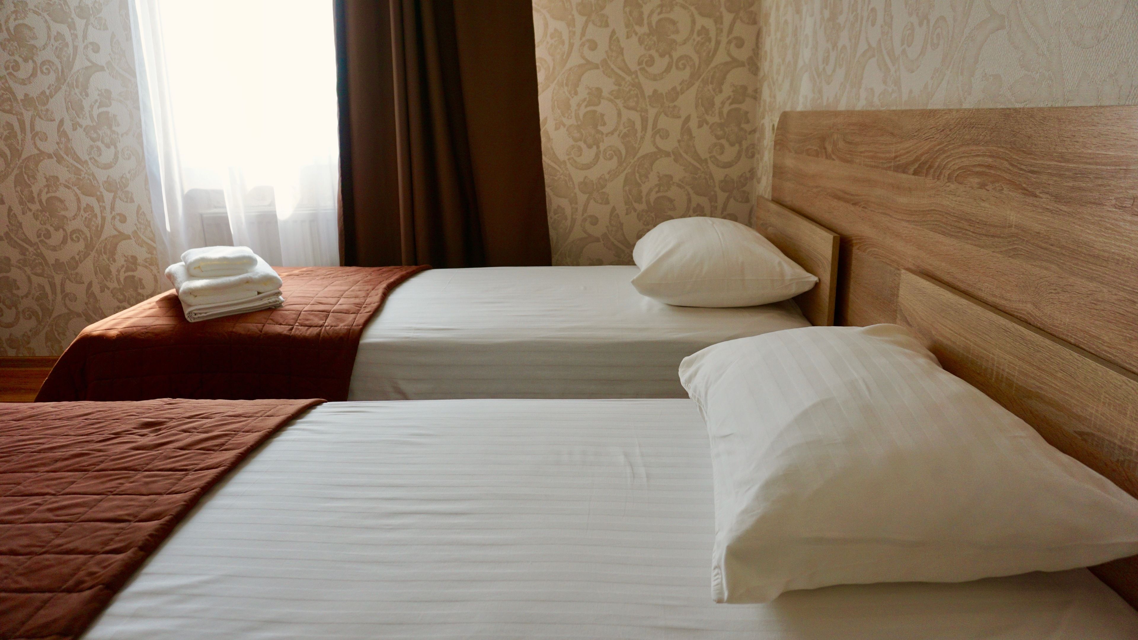 Ett hotellrum med två sängar, var och en med en sänggavel i trä och vitt sänglinne. En brun, vadderad sängkappa ligger på den vänstra sängen, och en vit kudde ligger på den högra sängen.