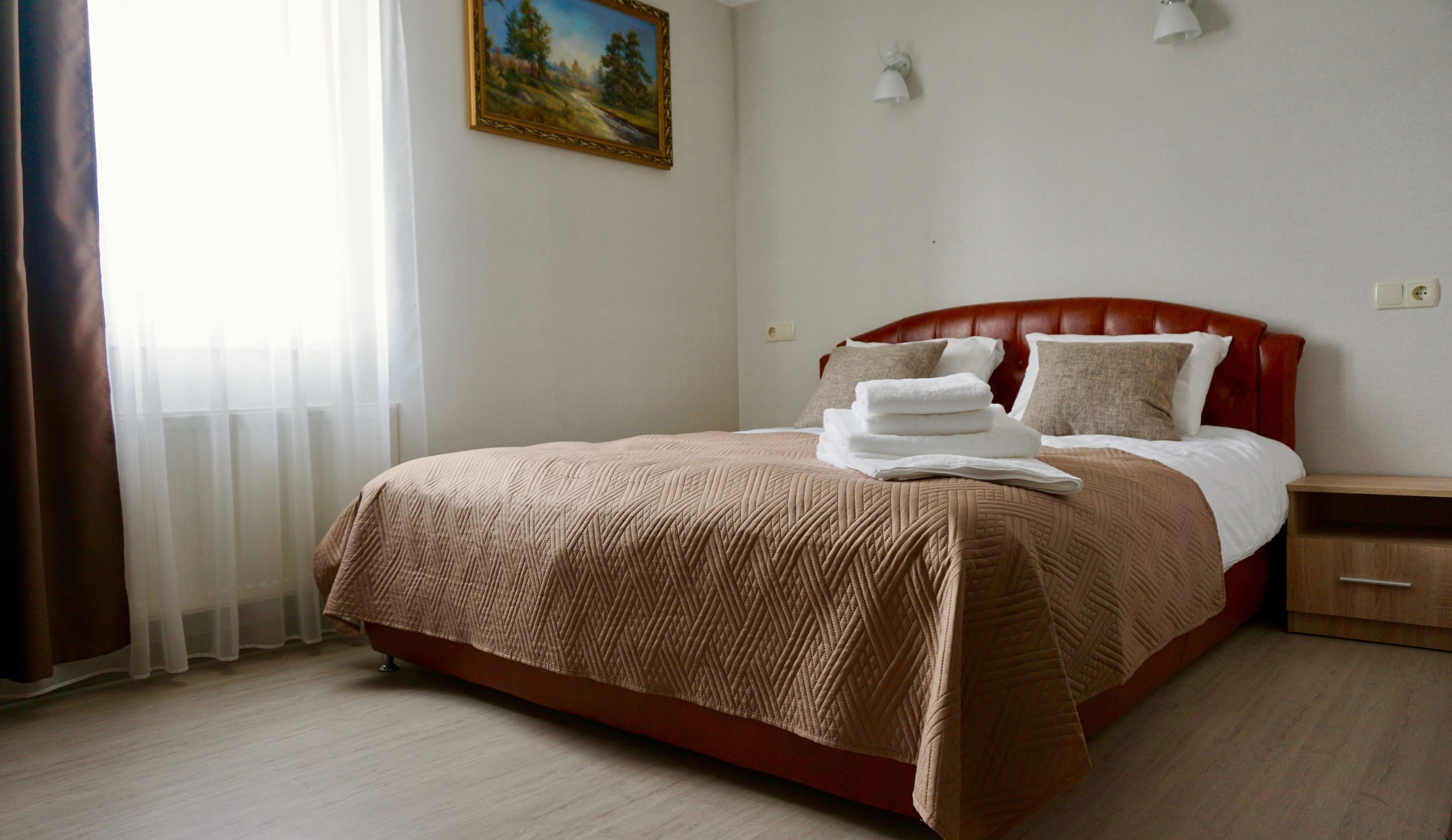A neatly made bed with a red headboard, white pillows, and a brown blanket. There is a painting on the wall above the bed and a nightstand with a lamp beside it.