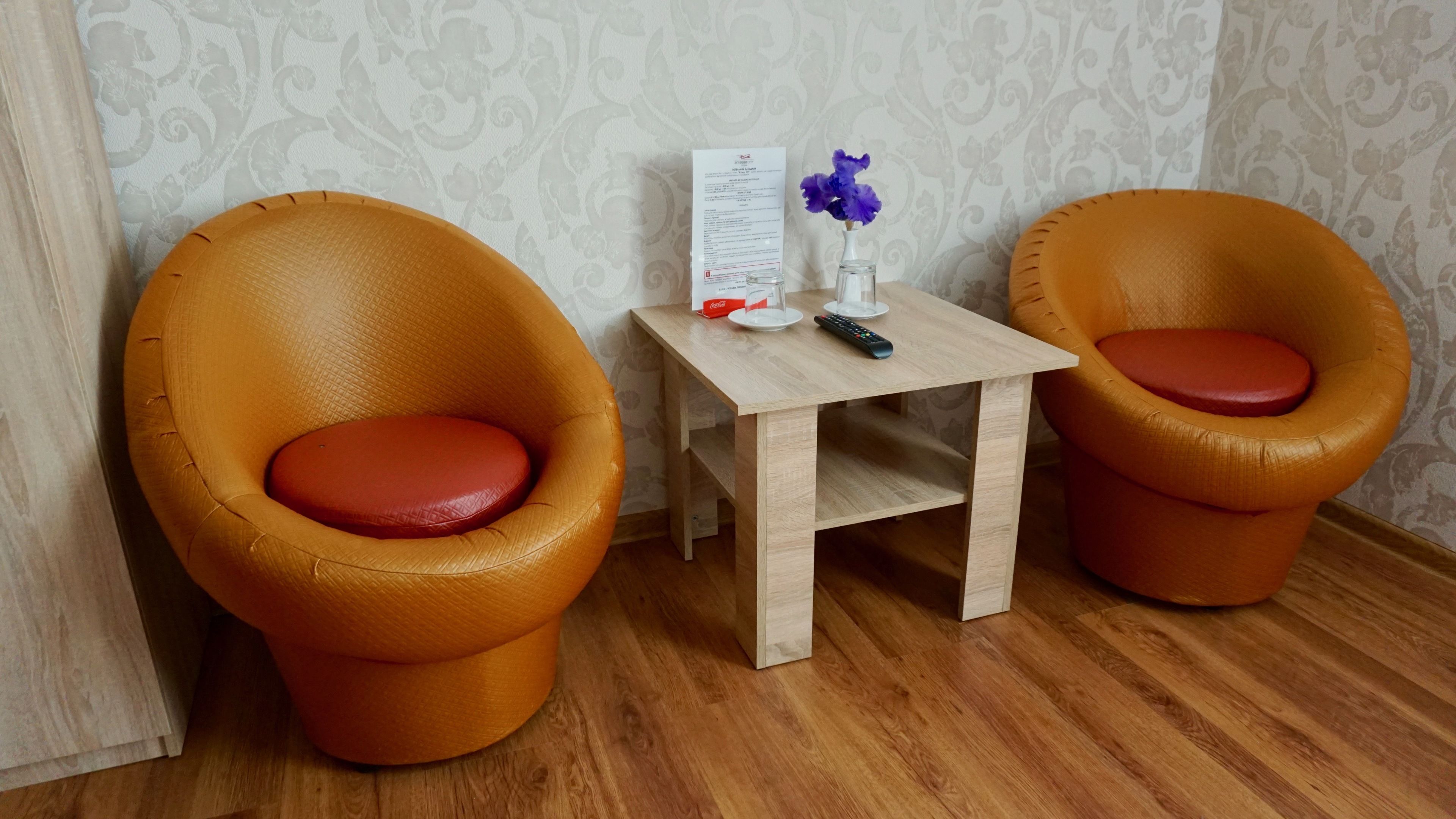 Two orange armchairs with red cushions, a small wooden table with a remote control, a vase with purple flowers, and a menu on the wall.