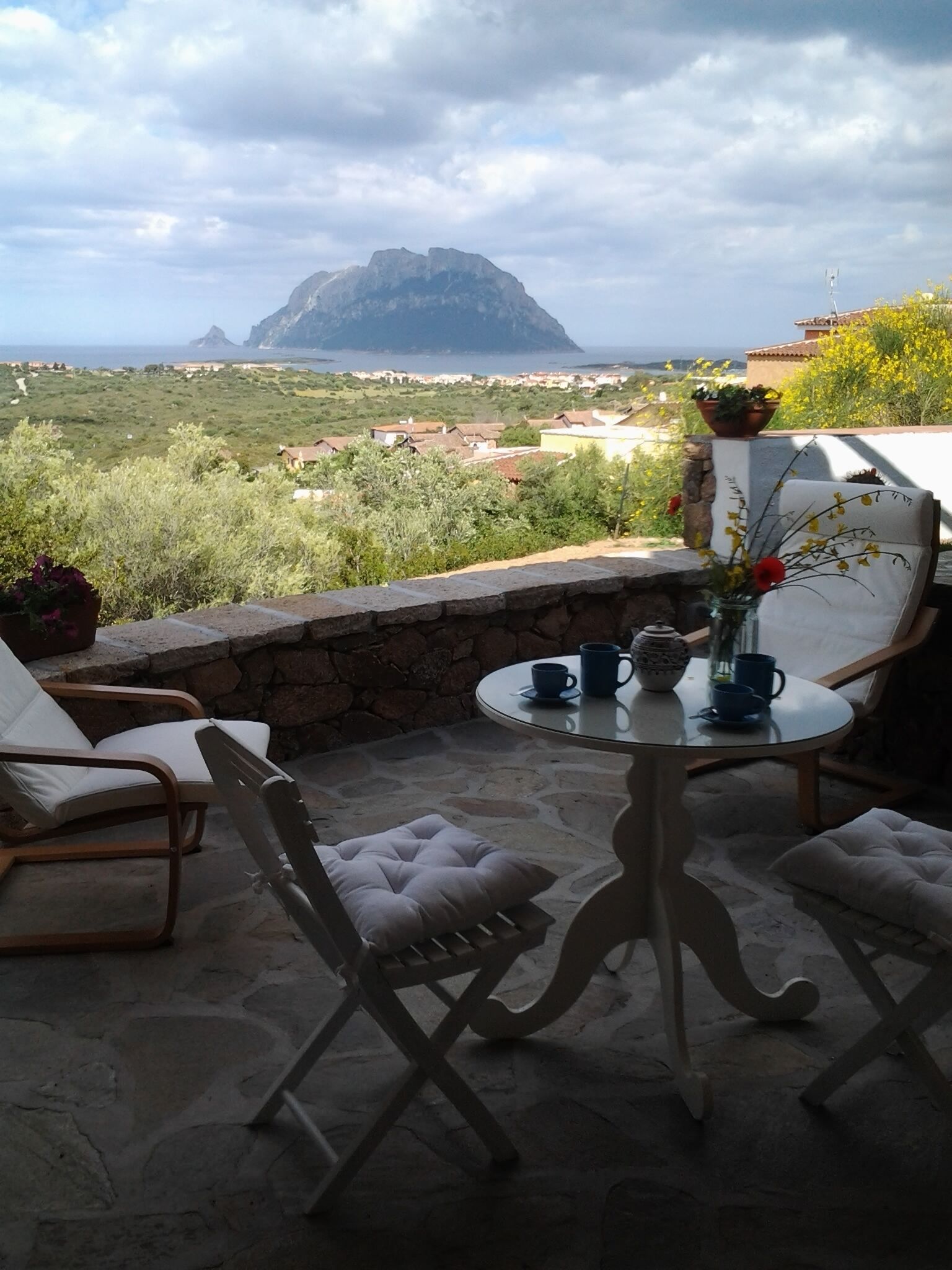 A terrace with a stone wall, two white cushioned chairs, a round table with a vase of flowers, and a view of a mountain and coastal town.