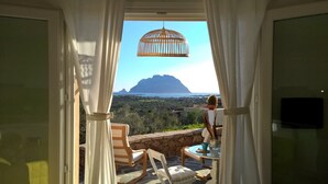 Un balcon offrant une vue sur la montagne et la mer, agrémenté d’un luminaire suspendu en osier et d’une table avec des chaises.