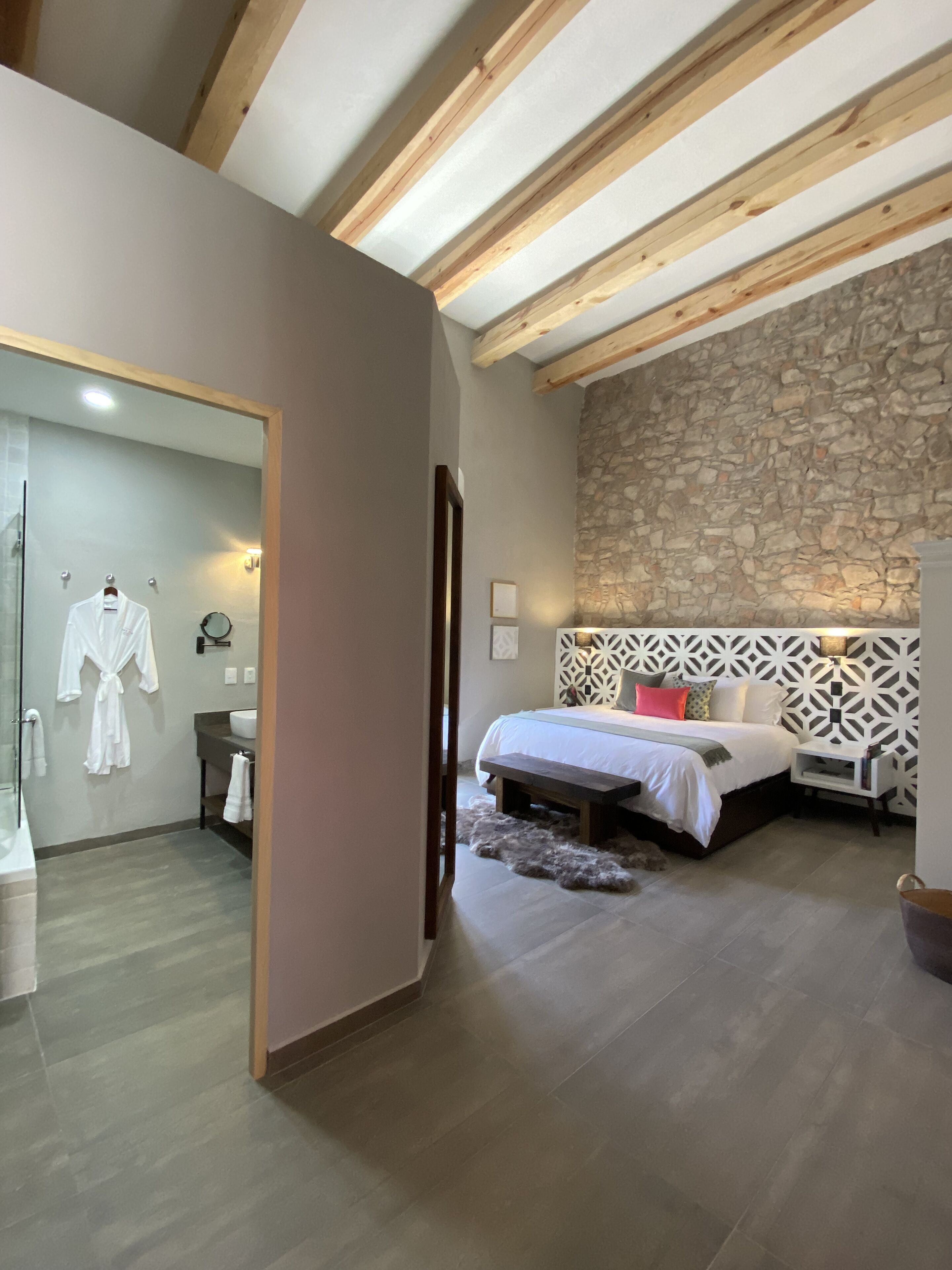 A bedroom with a stone wall, wooden beams, a bed, bedside table, and a bathroom with a mirror and robe.