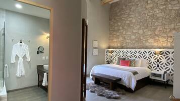 A bedroom with a stone wall, wooden beams, a bed, bedside table, and a bathroom with a mirror and robe.