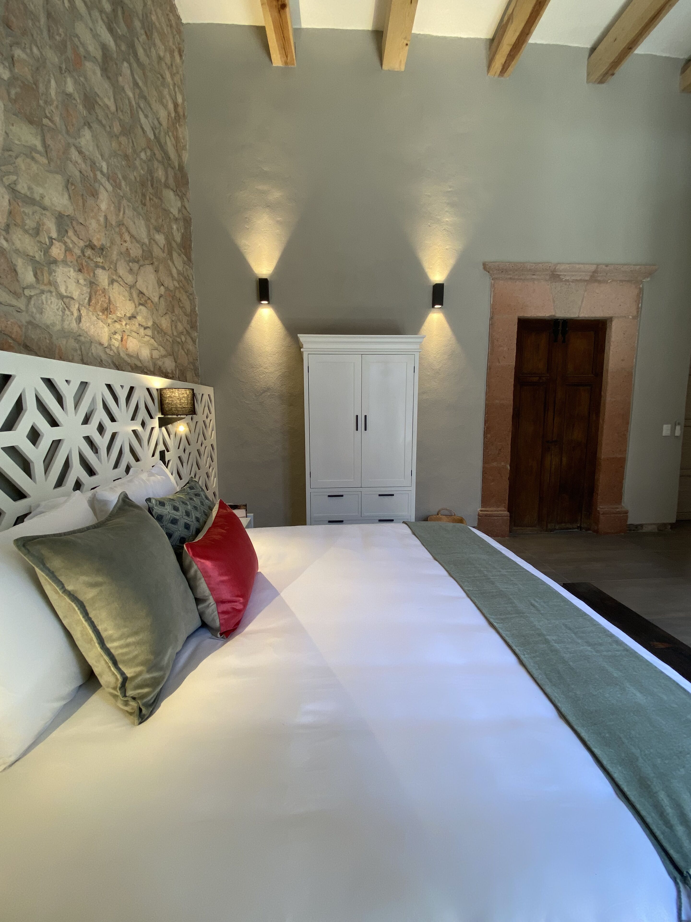 Un dormitorio con pared de piedra, cabecero blanco, cama con ropa de cama blanca, armario blanco y dos lámparas fijadas a la pared.