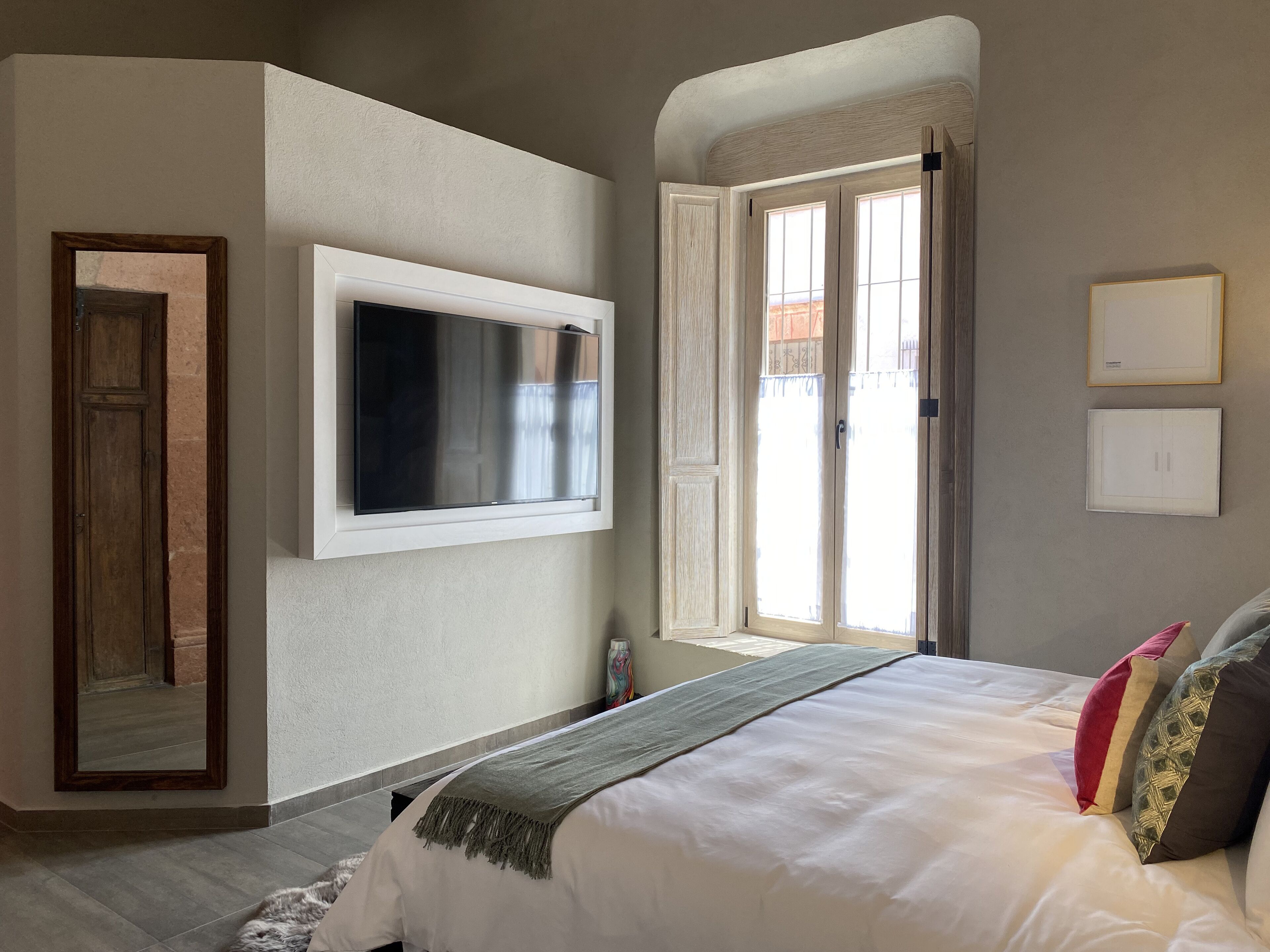 Un dormitorio con cama, televisor montado en la pared, una puerta y una ventana con cortinas.