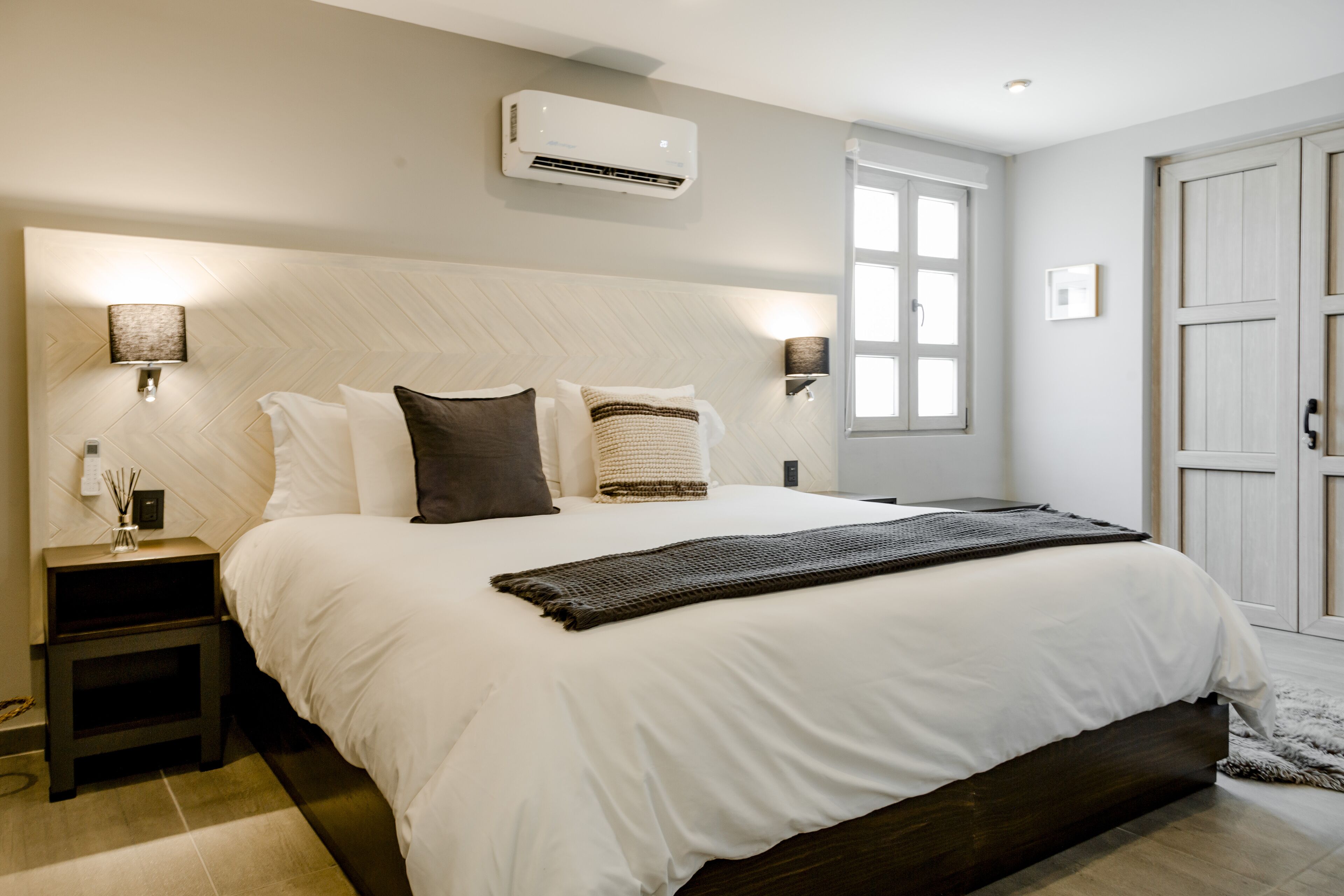 A modern bedroom with a large bed, a nightstand, a wall-mounted air conditioner, and a window with a view.