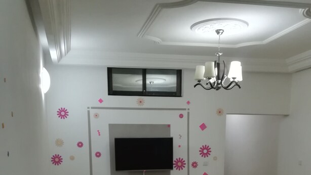 A living room with a large sofa, a flat-screen TV mounted on the wall, a chandelier, and a small table with a tissue box. - PRETTY T3 EQUIPPED COCODY (Cocody)