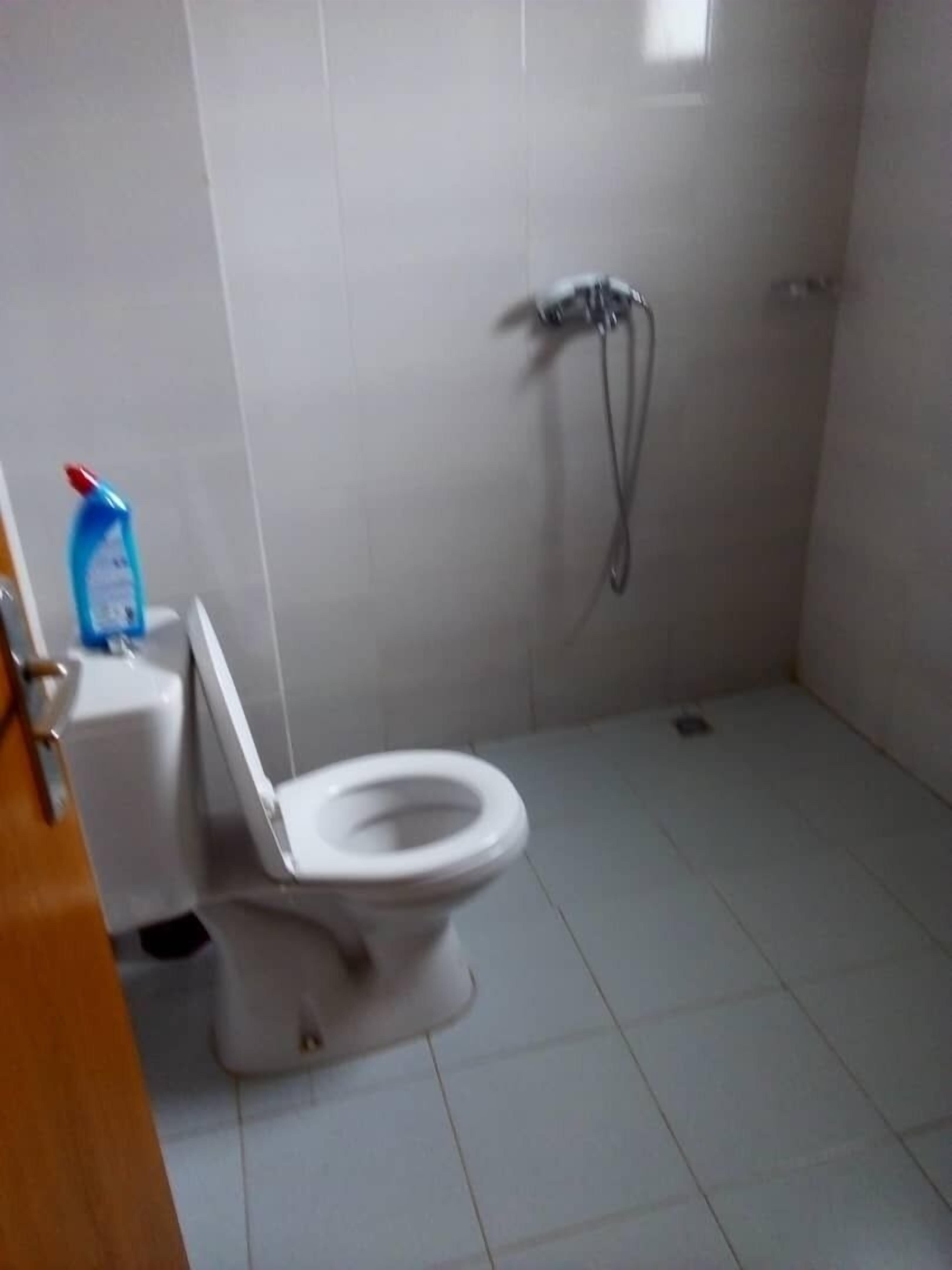 A small, tiled bathroom with a toilet, a shower, and a blue bottle of cleaning product on the floor.