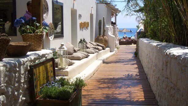 Een smal houten pad dat naar een strand leidt, begrensd door witte muren met decoratieve details en planten.