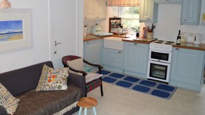 Une cuisine compacte avec des meubles de couleur bleu clair, une cuisinière et un évier. On y trouve un petit tabouret et une chaise avec un coussin à carreaux. Un tableau encadré est accroché au mur.