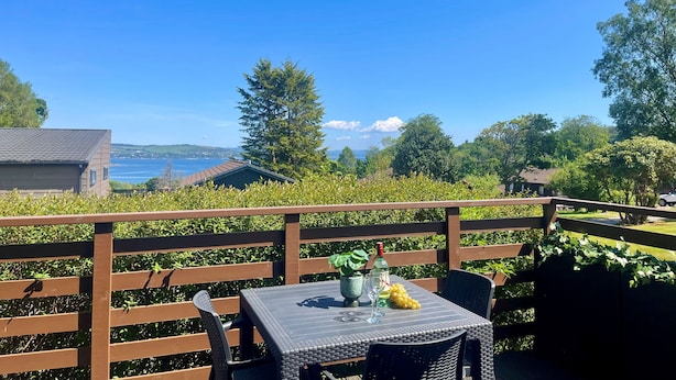 An outdoor patio with a table and chairs, overlooking a scenic view of a lake and distant buildings. - Loch view location in Holiday park, sleeps 4 persons, off road parking, free Wi-Fi (Dunoon)