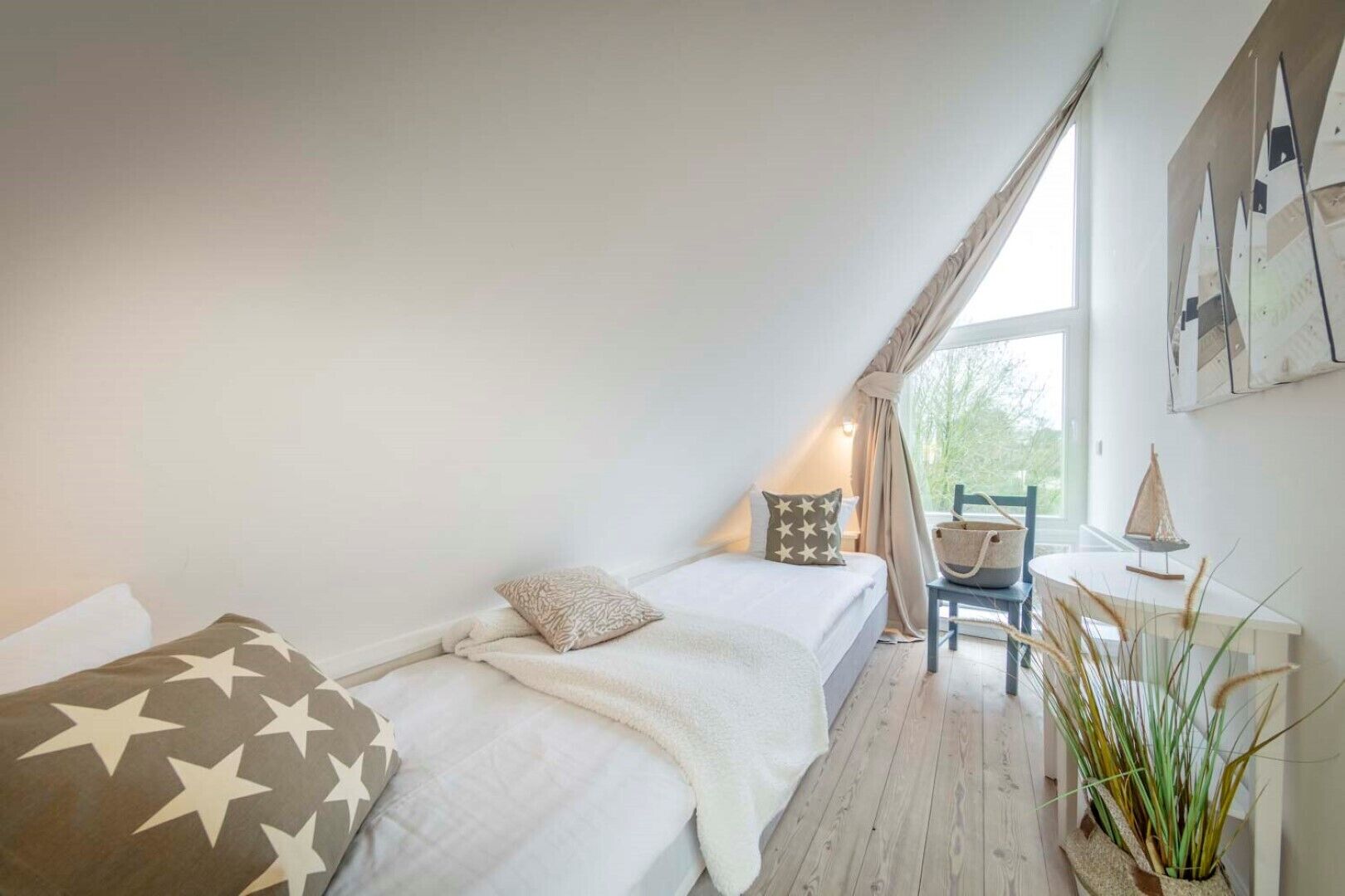 A cozy, well-lit attic bedroom with a sloped ceiling, a bed with star-patterned pillows, a small table with a lamp, and a view of greenery outside.