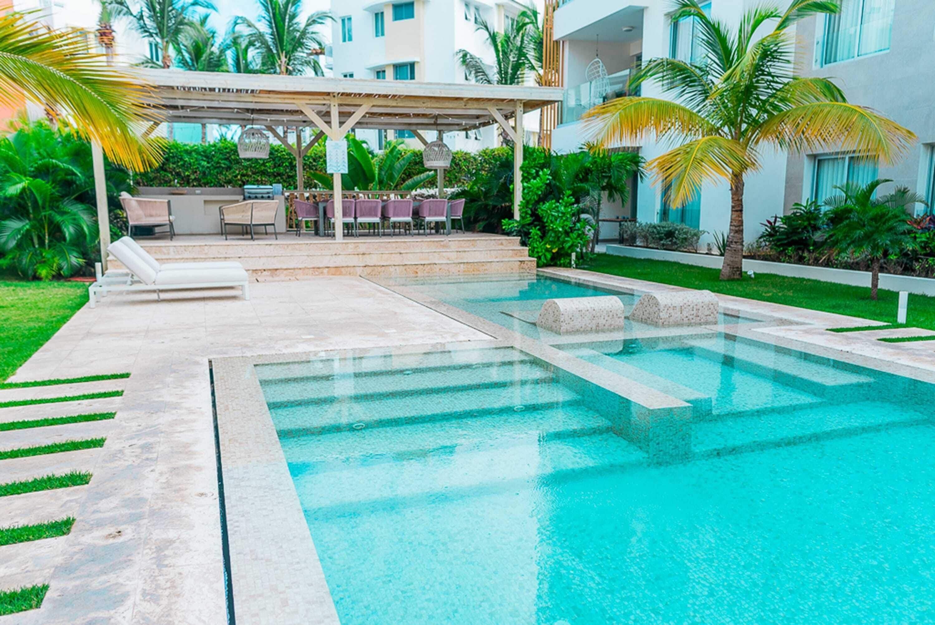 A swimming pool with a covered seating area, surrounded by greenery and palm trees.