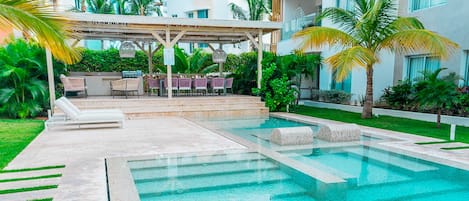 A swimming pool with a covered seating area, surrounded by greenery and palm trees.