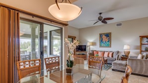 Un salon spacieux avec une table à manger en verre, des meubles en osier, un ventilateur de plafond et une grande fenêtre donnant sur l’extérieur.