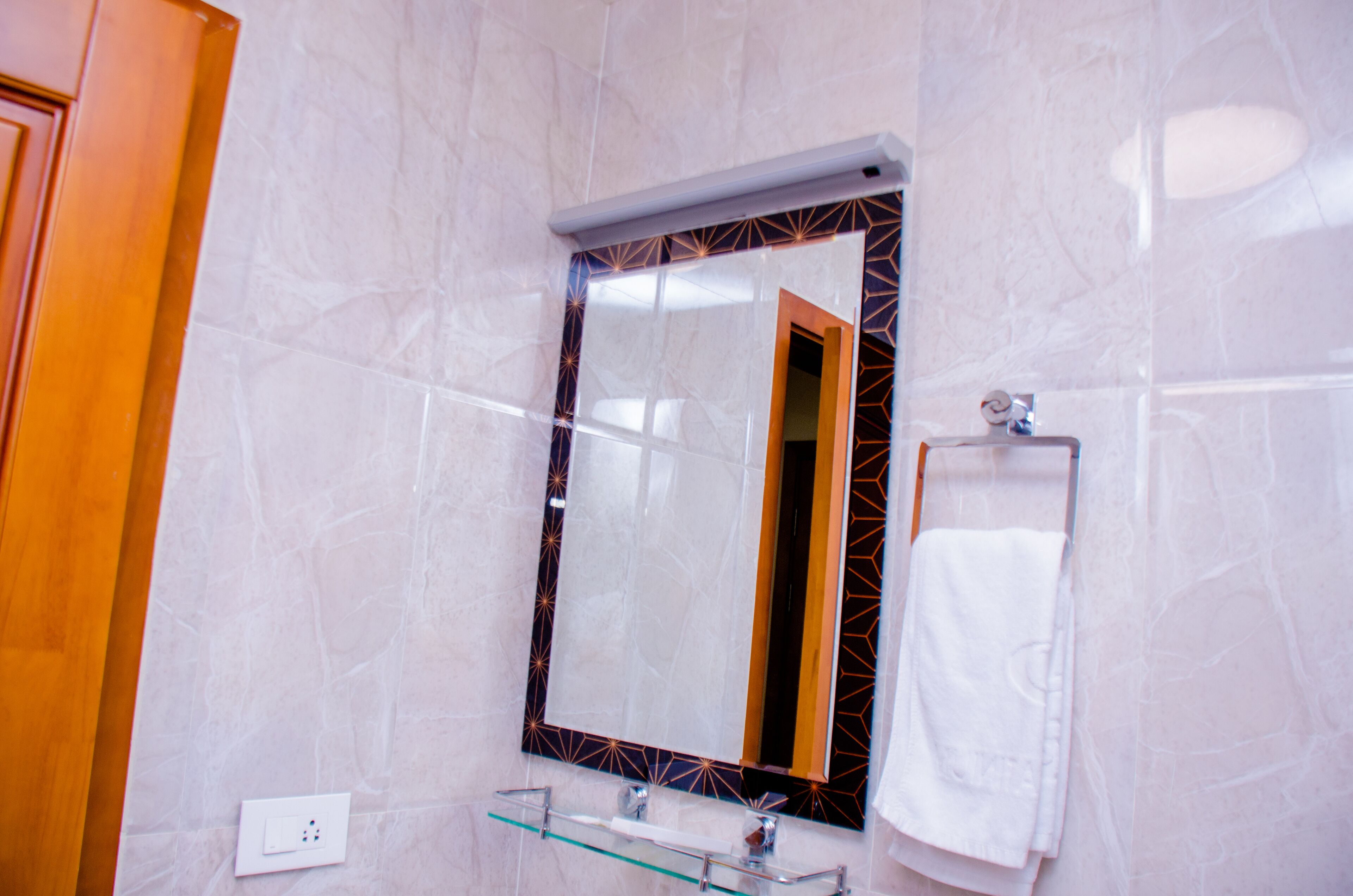 Une salle de bain avec un mur en marbre, une étagère en verre, une serviette et un miroir doté d’un cadre géométrique.