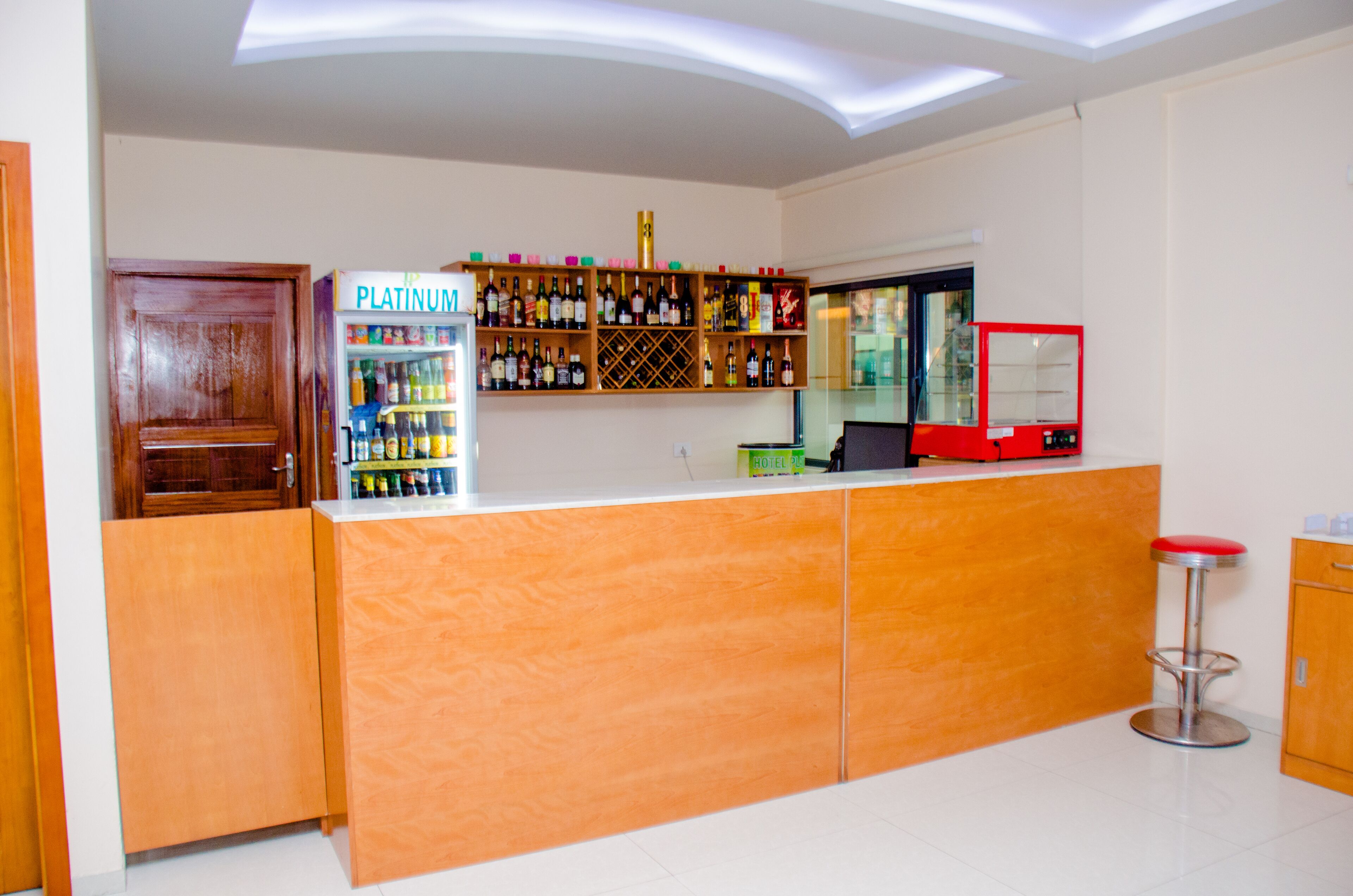 A hotel reception area with a wooden reception desk, a minibar, a red-framed glass display case, and a wooden door.