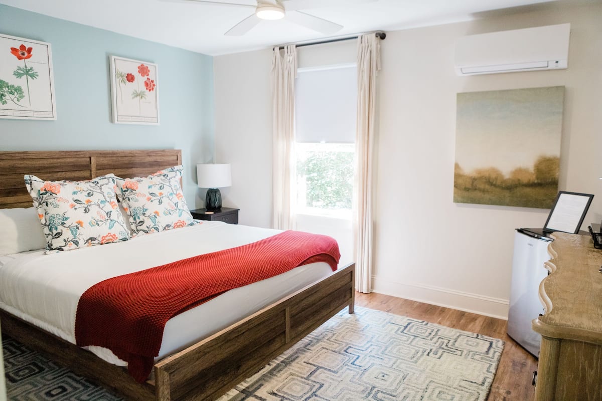 A bedroom with a wooden bed, floral pillows, a red blanket, a nightstand with a lamp, a wall-mounted air conditioner, and a large window with curtains.