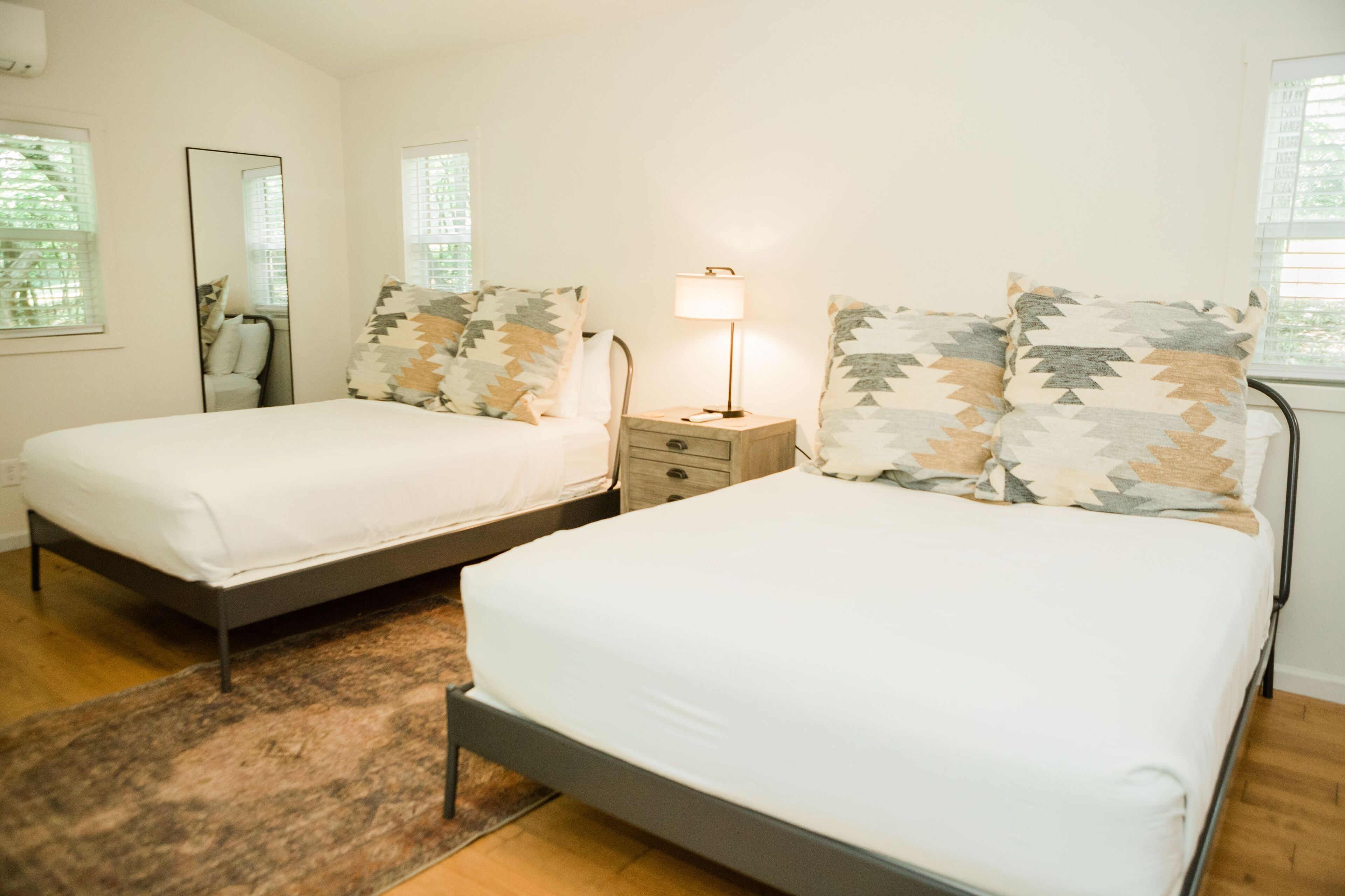 A double bed with a white mattress and patterned pillows, a bedside table with a lamp, and a mirror on the wall.