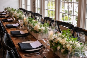 Uma longa mesa de madeira posta para um evento formal, com cadeiras, copos e arranjos florais.