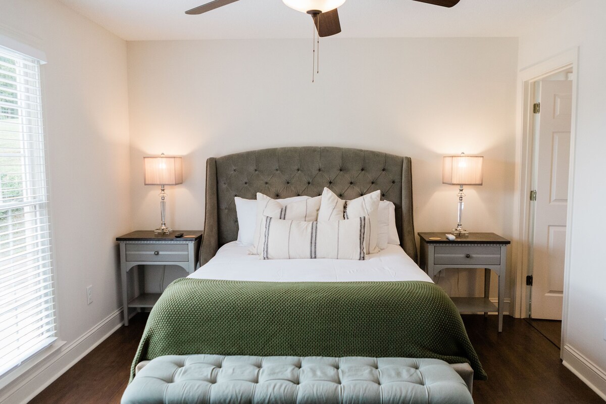 A bedroom with a tufted headboard, a green bedspread, two bedside tables with lamps, and a ceiling fan.
