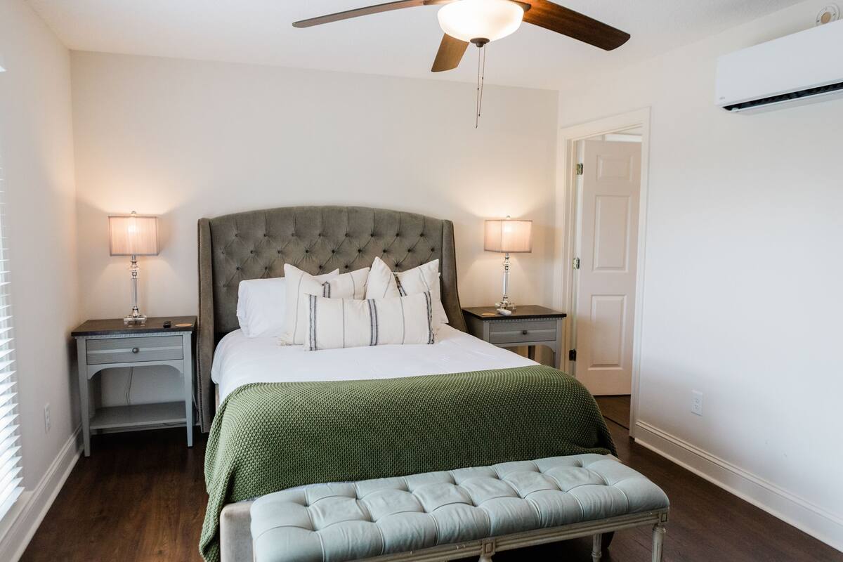 A bedroom with a tufted headboard, a green bedspread, two bedside lamps, a bench, and a ceiling fan.