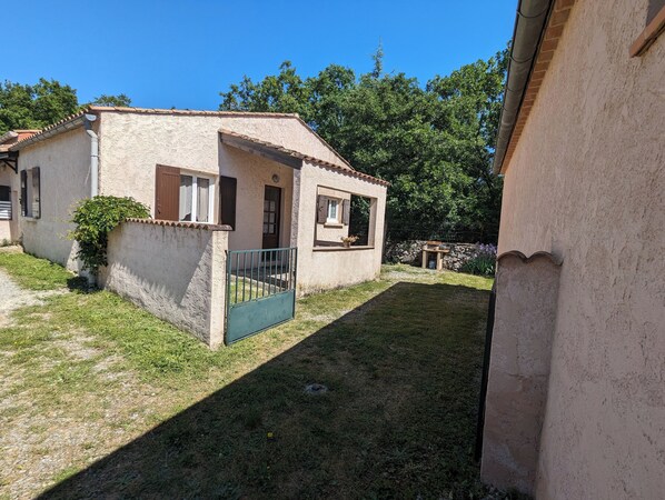 Une petite maison à un étage, avec un toit en tuiles, une cour clôturée et un mur de pierre en arrière-plan.