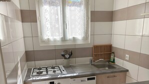 A compact kitchen with a gas stove, oven, dishwasher, and a window with lace curtains.