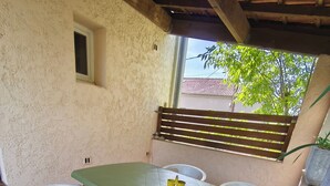Une terrasse avec une table verte et des chaises en plastique blanches, une fenêtre donnant sur des arbres et une clôture en bois.