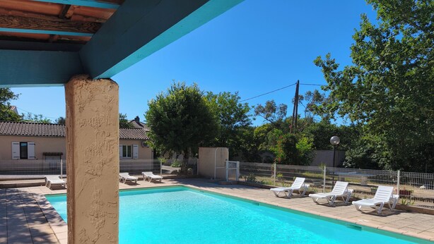 Une piscine avec des chaises longues, une terrasse carrelée et un espace clos, agrémenté d’arbres et avec un bâtiment en arrière-plan.