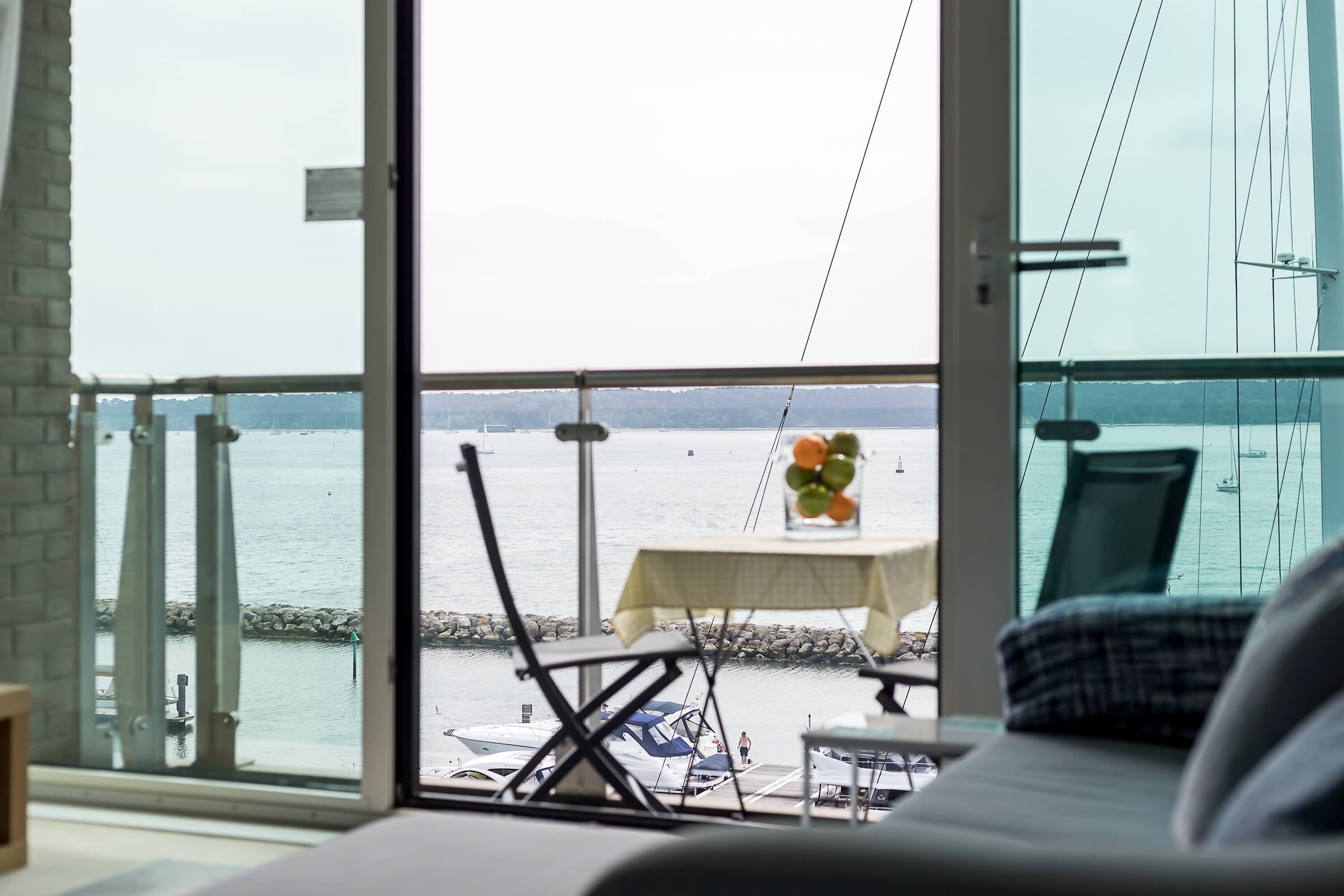 A room with a large window overlooking a marina with boats. There is a table with a vase of flowers and a chair.