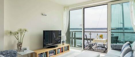 A modern living room with a flat-screen TV, bookshelves, a sofa, and a balcony with a view of the sea.
