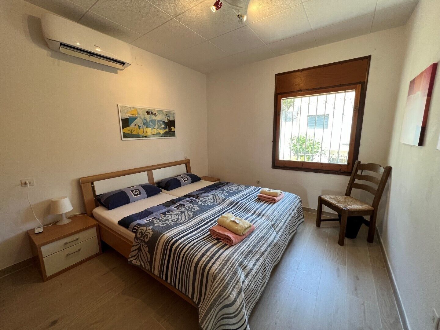 A bedroom with a bed, bedside table, lamp, chair, window, and artwork on the wall.