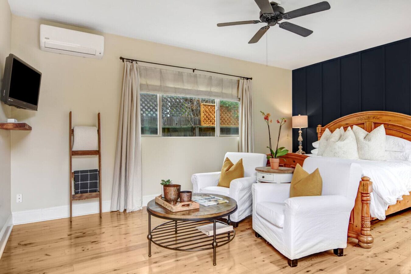 A bedroom with a bed, two white armchairs, a small round table, a wooden ladder, a ceiling fan, and a window with curtains.