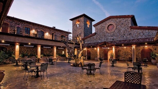 Un patio con edificios de piedra, asientos al aire libre y un árbol en el centro.
