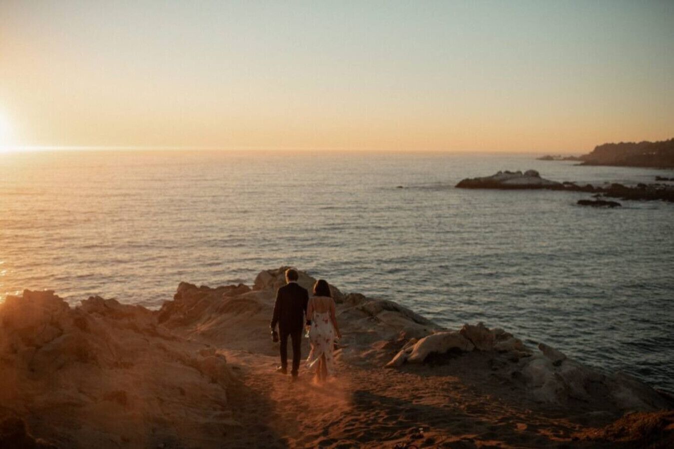 Una pareja camina sobre un sendero rocoso junto al mar durante el atardecer.