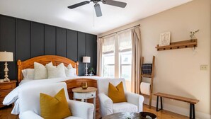 Habitación de hotel con una cama grande, dos sillones blancos con cojines mostaza, una pequeña mesa redonda, un ventilador de techo, una ventana con cortinas y una estantería de madera con una planta.