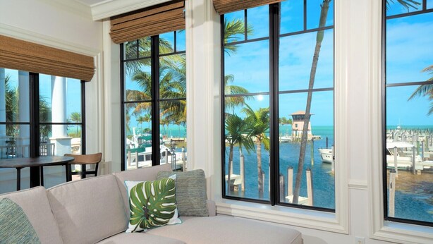 Una sala de estar moderna con un sofá seccional color beige, ventanas grandes con persianas de madera y vista a un puerto deportivo y una playa.