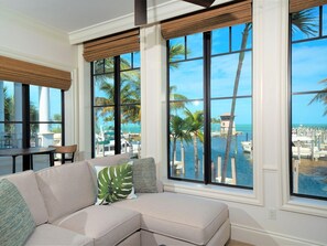 Una sala moderna con un sofá seccional color beige, ventanas amplias con persianas de bambú y vista a un puerto deportivo con barcos y palmeras.