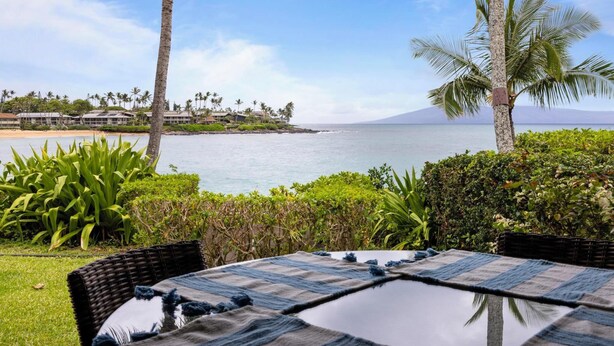 A view of a beachside property with a table and chairs, palm trees, and a distant view of a mountain. - Napili Lani 315  2BD Beachfront Luxury (Lahaina)