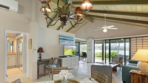 A spacious living area with a dining table, chairs, a television, and a large window offering a view of the outdoors. - Napili Lani 315  2BD Beachfront Luxury (Lahaina)