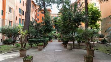 A courtyard with a paved walkway, surrounded by trees and potted plants, leading to a building with multiple windows.