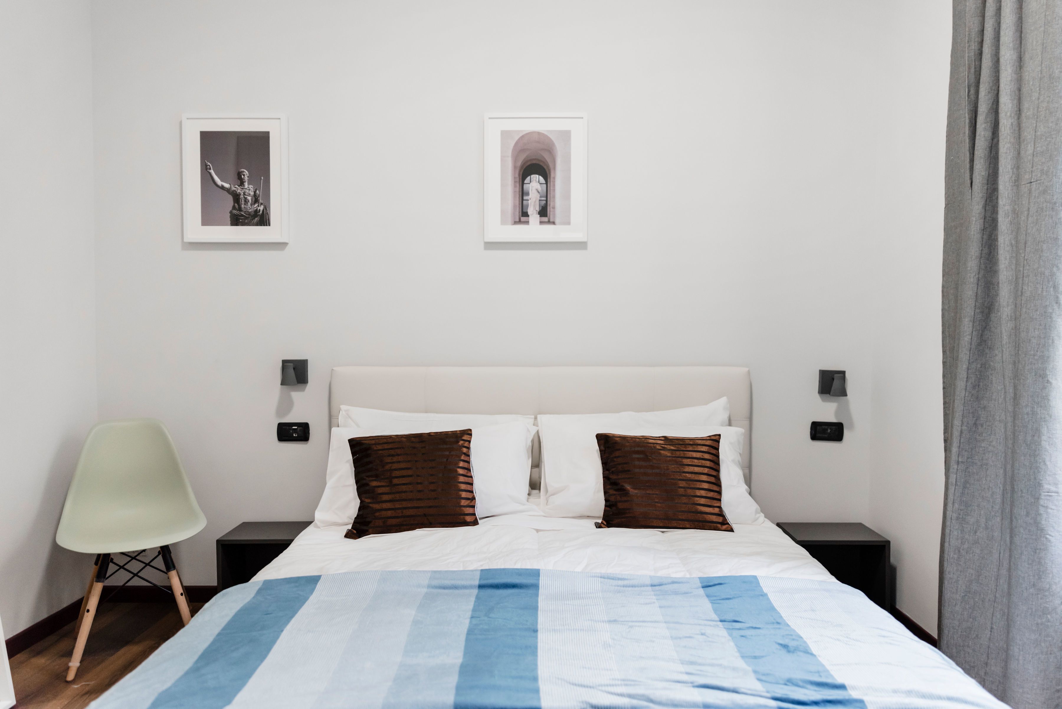 A hotel room with a bed, two pillows, a nightstand, a chair, and two framed pictures on the wall.