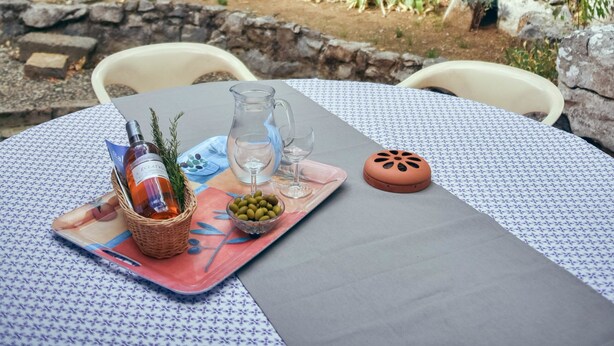An outdoor dining area with a table set for two, featuring a bottle, olives, and a pitcher. There are two green chairs and a stone wall in the background.