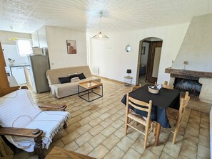 A living room with a sofa, a dining table with chairs, a fireplace, and a kitchen area in the background.