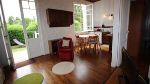 Uma sala de estar espaçosa com piso de madeira, uma mesa de jantar com cadeiras, uma poltrona vermelha e uma grande janela com vista para o verde lá fora.