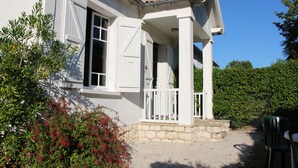 Uma casa branca com alicerce de pedra, varanda com grades brancas e um pequeno jardim com flores vermelhas.