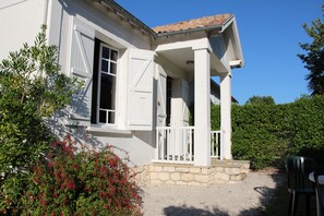Ein weißes Haus mit Stein Fundament, einer Veranda mit weißen Geländern und einem kleinen Garten mit roten Blumen.