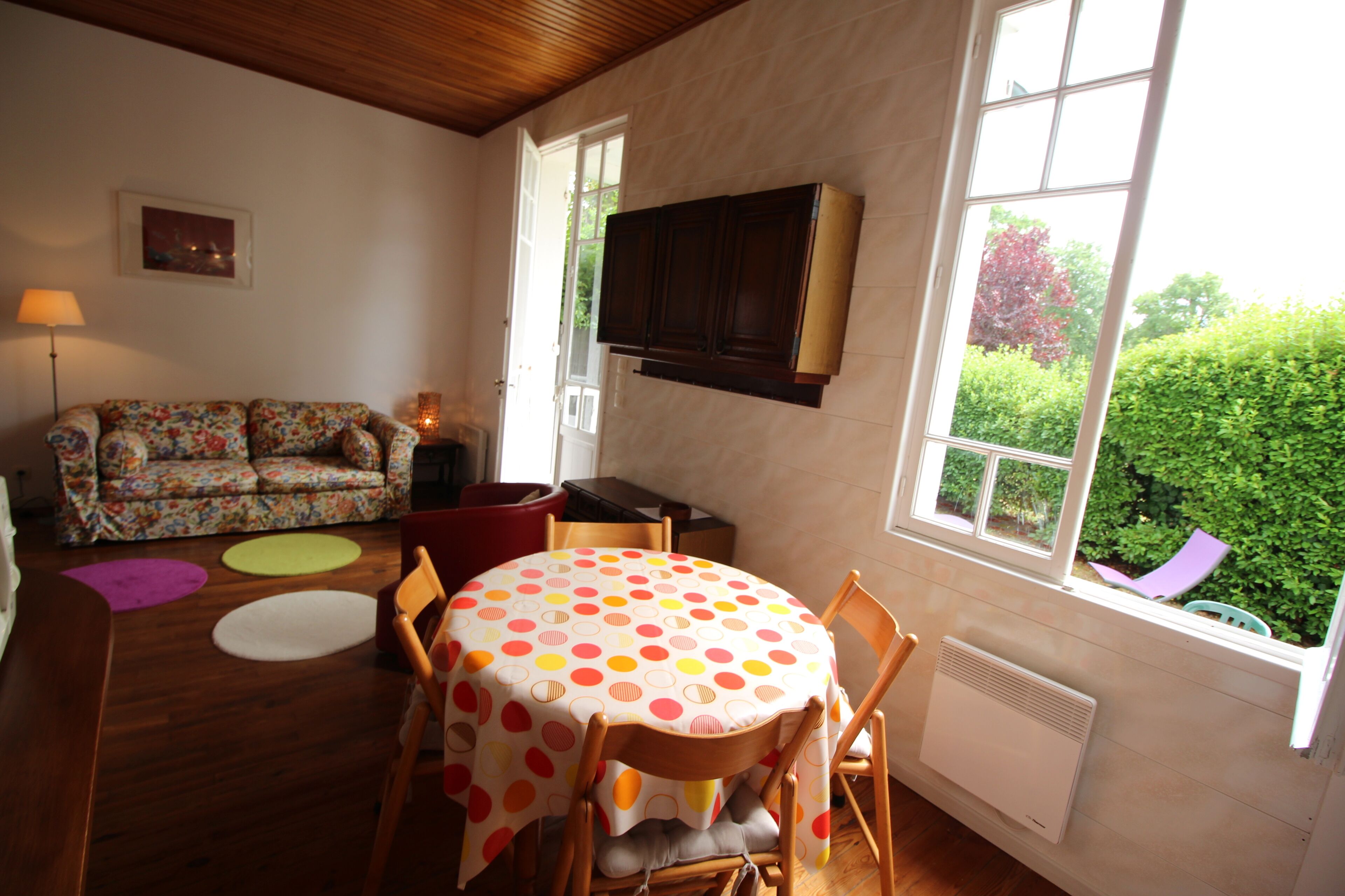 A dining area with a round table covered by a polka dot tablecloth, wooden chairs, and a floral sofa. A window provides a view of greenery outside.