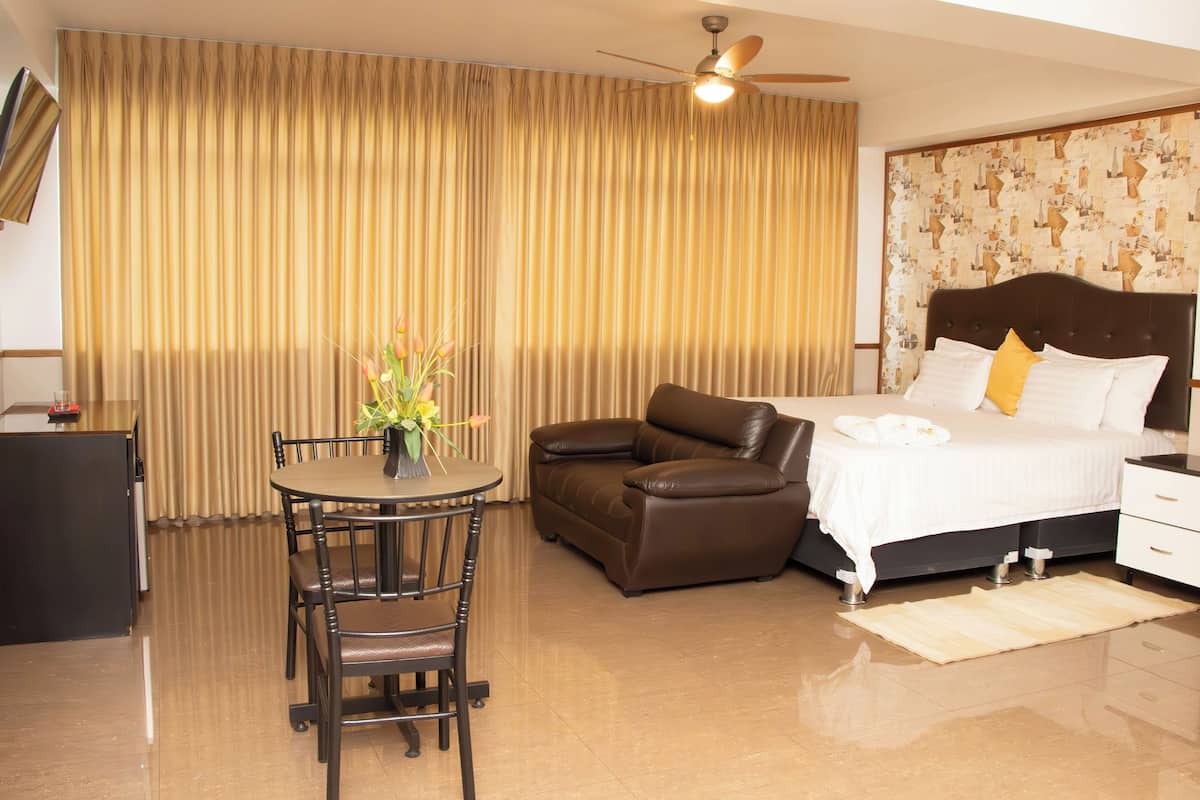 A hotel room with a bed, a brown leather sofa, a dining table with chairs, a small desk, and a television.